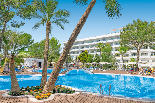 Poolanlage eines Hotels in ovaler Form mit Palmen, Liegen und im Hintergrund das weiße Hotelgebäude